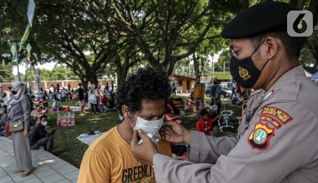 Polisi memasangkan masker untuk pengunjung yang berwisata di Taman Mini Indonesia Indah (TMII), Jakarta, Sabtu (15/5/2021). Selama libur Lebaran, pengelola TMII memberlakukan protokol kesehatan secara ketat. (Liputan6.com/Faizal Fanani)