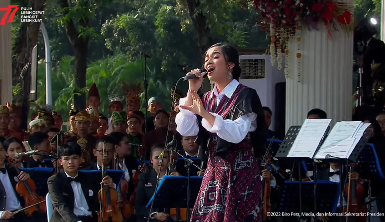 Sementara itu, Lyodra tampil cantik mengenakan outfit dengan sentuhan kain tenun. Memilih kemeja putih berlengan mangkuk sebagai inner dipadukan dengan outer menyilang dari kain tenun. (Tangkapan layar YouTube SetPres RI)