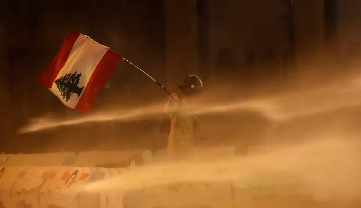 Demonstran antipemerintah mengibarkan bendera Lebanon saat dia disemprot air dalam demonstrasi menentang pemerintahan baru dekat Parliament Square, Beirut, Lebanon, Rabu (22/1/2020). Demonstrasi dan kekerasan terus berlanjut kendati Lebanon telah mengumumkan kabinet baru. (AP Photo/Hussein Malla)