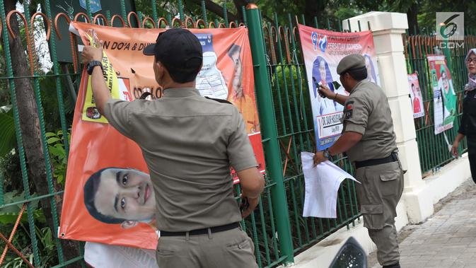 Petugas Satpol PP bersama Panwaslu kecamatan Ciracas menurunkan APK yang terpasang di pagar pembatas antara jalan dengan taman di pinggir kali di sekitar Jalan Raya Bogor, Jakarta, Rabu (23/1). (Liputan6.com/Helmi Fithriansyah)