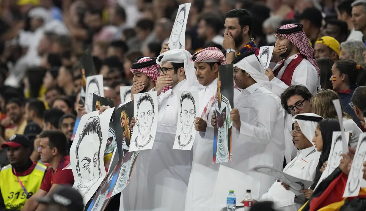 Penonton memegang sketsa Mesut Ozil di tribun selama pertandingan Spanyol vs Jerman pada laga Grup E Piala Dunia 2022  di Stadion Al Bayt, Kota Al Khor, Senin (28/11/2022) dini hari WIB. Aksi sindiran balik dari tribune Al-Bayt itu kemudian merujuk kepada tindakan rasialisme yang dilakukan Jerman kepada mantan pemainnya tersebut.  (AP Photo/Ebrahim Noroozi)