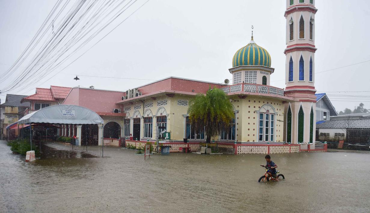 Setidaknya 13 orang dilaporkan tewas akibat banjir di beberapa provinsi di Thailand selatan, termasuk Songkhla. Tampak dalam foto, seorang anak bersepeda di tengah banjir di dekat sebuah masjid di provinsi Narathiwat, Thailand selatan, pada Selasa 25 November 2025. (Madaree TOHLALA/AFP)