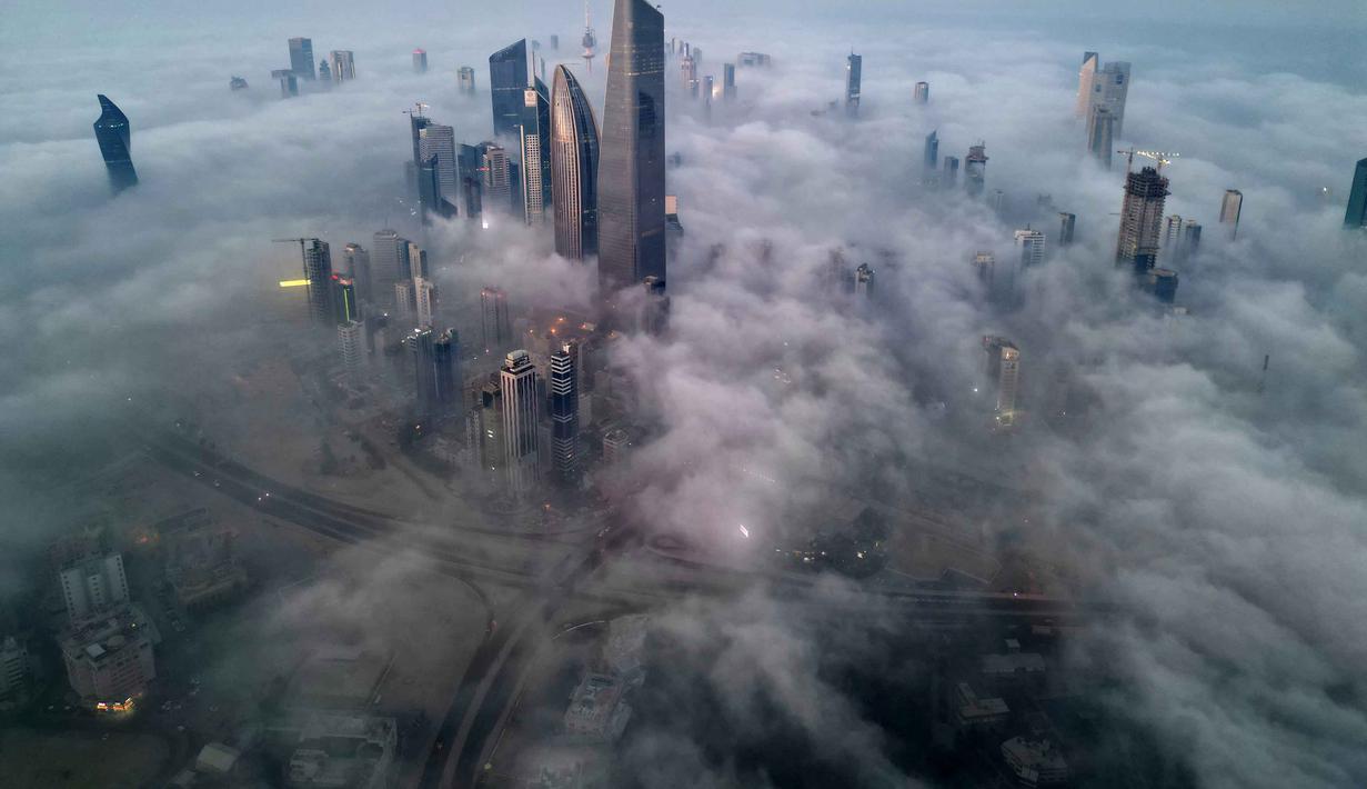 Pemandangan ini menciptakan ilusi kota di atas awan, dengan bagian atas gedung menembus kabut tebal. Tampak foto udara memperlihatkan gedung pencakar langit yang tertutup kabut pagi di Kota Kuwait pada Minggu 9 November 2025. (YASSER AL-ZAYYAT/AFP)