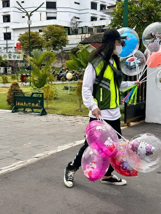 Dalam potret yang dibagikan, selain tangan kanan dan kirinya memegang balon, Baim juga dandan ala kadarnya. Memakai sepatu, rompi, dengan rambut panjang. Baim juga mengenakan topi hitam dan masker putih. [Instagram/baimwong]