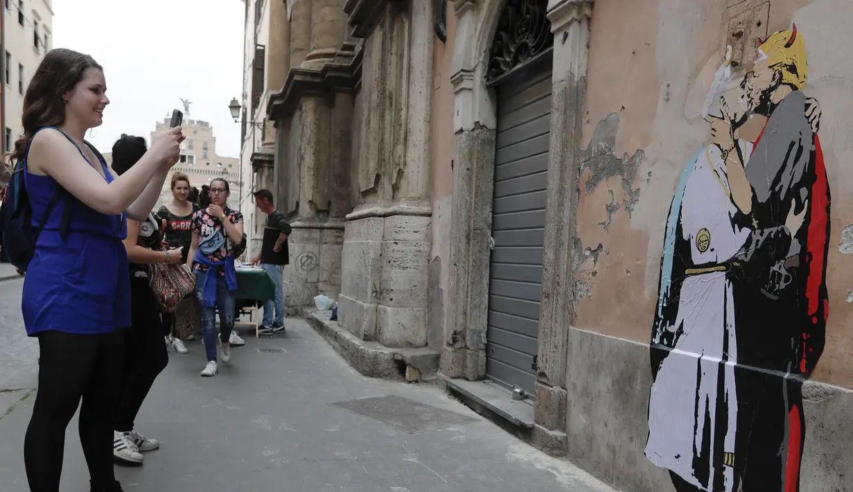 Pejalan kaki memotret mural Presiden AS, Donald Trump dan Paus Francis berciuman di sebuah dinding di Roma, Kamis (11/5). Dalam mural itu Fransiskus berpakaian putih dan memeluk Trump yang memiliki tanduk merah dan pistol. (AP Photo/Alessandra Tarantino)