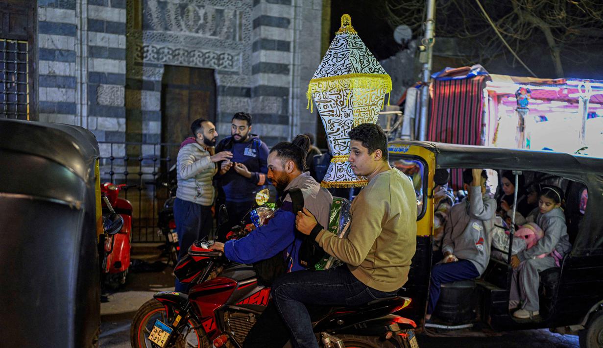 Fanous bukan hanya dekorasi, tetapi simbol budaya yang memperindah suasana malam Ramadan di Mesir. Tampak dalam foto, seorang pria Mesir mengendarai sepeda motor membawa lentera tradisional, yang dikenal secara lokal sebagai 'Fanous Ramadan', di kawasan kota tua Kairo pada Kamis 12 Februari 2026. (Khaled DESOUKI/AFP)