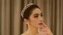 Gaun pengantin yang dikenakan Angel Karamoy memiliki potongan square neckline yang elegan dengan lengan panjang berbahan lace transparan. Siluet ball gown yang mengembang dengan ekor panjang menambah kesan mewah bak putri kerajaan. Aksesori tiara kristal dan anting mutiara semakin mempercantik tampilannya. (Liputan6.com/IG/@realangelkaramoy)