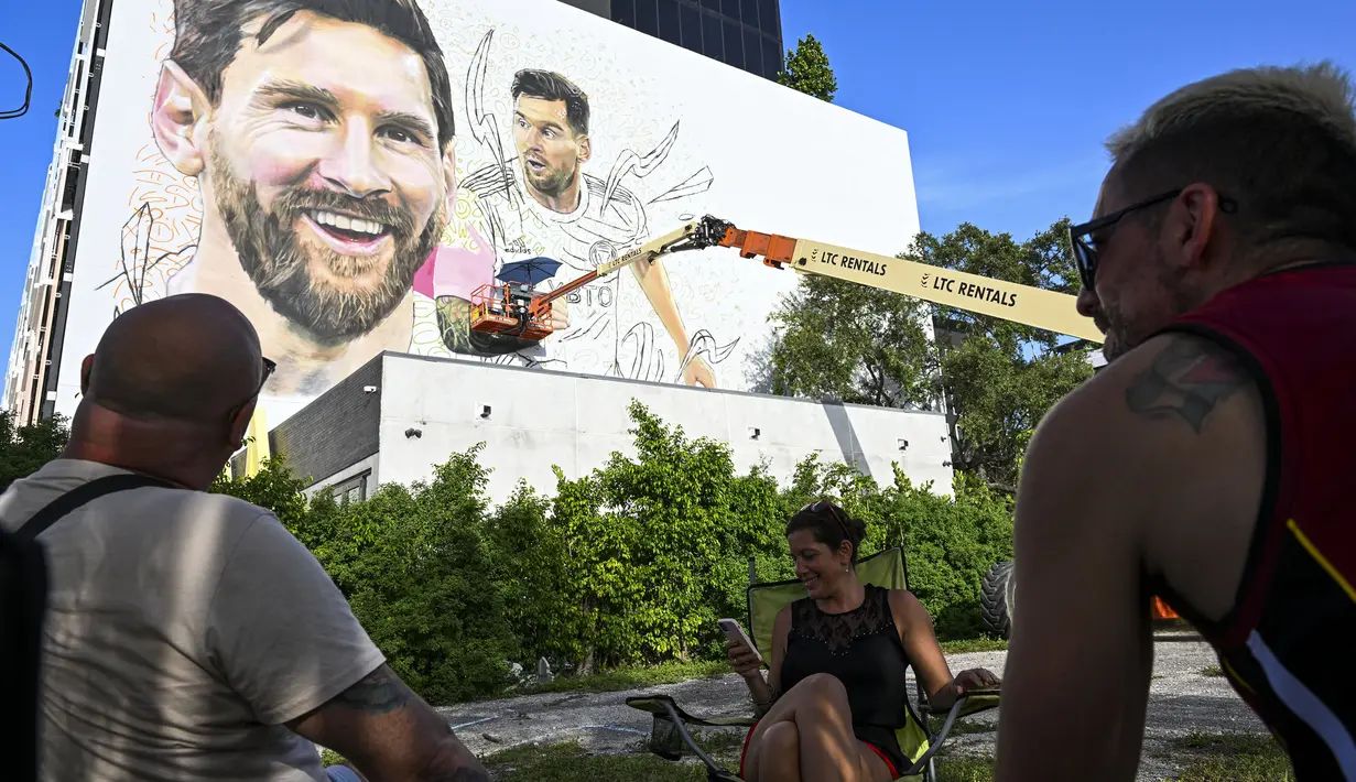 <p>Sejumlah warga menonton seniman Argentina, Maximiliano Bagnasco saat mengerjakan mural besar bergambar Lionel Messi di Wynwood, Miami, 10 Juli 2023. La Pulga akan melakukan debutnya di Inter Miami pada 21 Juli di Piala Liga. (AFP/Chandan Khanna)</p>