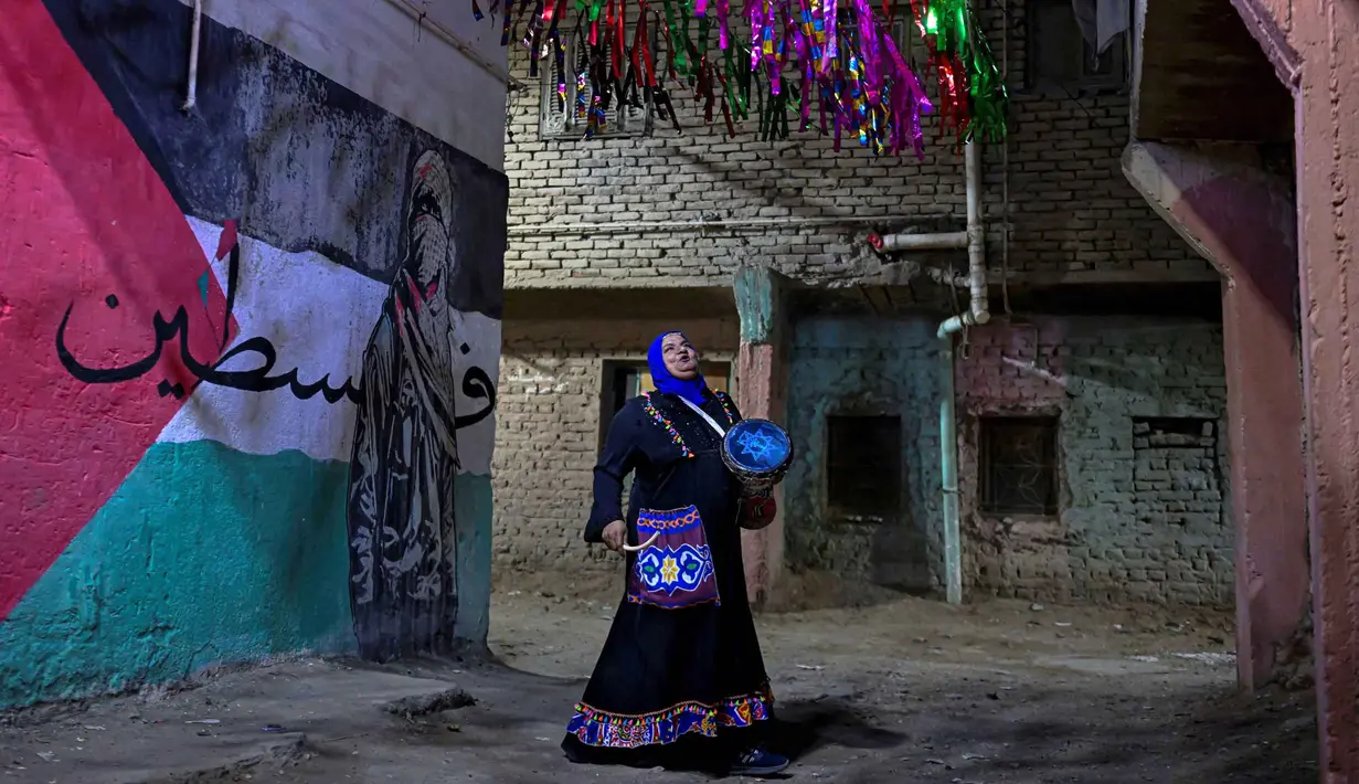 Dulu, petugas ini ditunjuk secara resmi untuk memastikan tidak ada warga yang melewatkan sahur. Tampak dalam foto, Dalal, seorang wanita Mesir 'musaharati', berdiri di depan grafiti yang menggambarkan bendera Palestina di sepanjang jalan sambil memukul gendang dan menyerukan umat Muslim untuk bangun dan makan sahur selama bulan suci Ramadan di distrik Hadayek el-Maadi, selatan Kairo pada Minggu 1 Maret 2026. (Khaled DESOUKI/AFP)