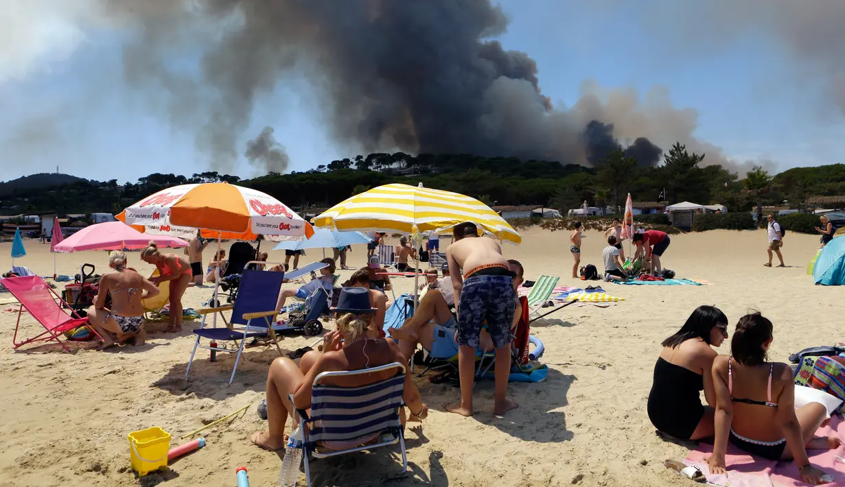 Wisatawan berjemur sambil menyaksikan asap hitam yang membumbung akibat kebakaran hutan dari pinggir pantai Lavandou, Rabu (26/7). Kebakaran menghanguskan hamparan hutan di Prancis selatan dan di Pulau Corsica dalam tiga hari terakhir. (AP/Claude Paris)