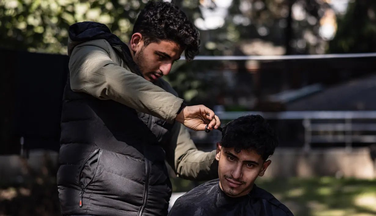 Mohammed al-Hamo (kiri) memotong rambut saudara laki-lakinya, Sobhi di depan tenda mereka di kamp darurat pengungsi korban gempa Turki dan Suriah di kota Antakya pada 19 Februari 2023. Hamo berhasil mengambil gunting listrik, sisir, gunting, dan sampo dari reruntuhan rumahnya yang hancur. (Sameer Al-DOUMY / AFP)