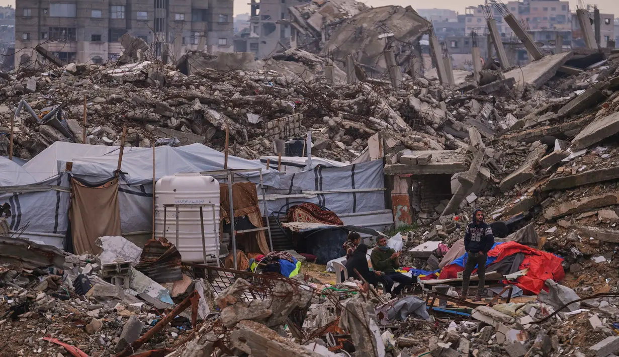 Warga Palestina berdiri di samping tenda yang didirikan di atas reruntuhan bangunan yang hancur akibat serangan udara dan darat Israel di lingkungan Sheikh Radwan, Kota Gaza, Selasa, 30 Desember 2025. Setelah lebih dari dua tahun agresi militer Israel yang menghancurkan wilayah tersebut, kini cuaca dingin ekstrem dan hujan lebat menjadi tantangan besar bagi pengungsi di Jalur Gaza. (AP Photo/Abdel Kareem Hana)
