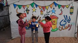 Beberapa anak tampak antusias menggantung hiasan sederhana di antara tali-tali penyangga tenda. Tampak dalam foto, para pengungsi muda Palestina bermain dengan lampion Ramadan saat dekorasi dipasang di samping tenda mereka di kamp pengungsi Bureij di Jalur Gaza tengah pada 15 Februari 2026. (Eyad Baba/AFP)