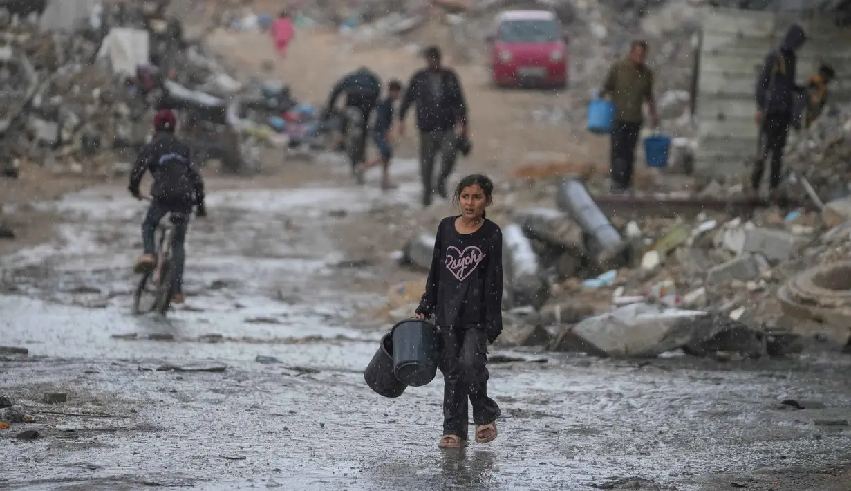 Seorang anak perempuan membawa dua ember kosong saat berjalan menuju tempat mengambil air di tengah hujan deras di kawasan Sheikh Radwan, Kota Gaza, Jumat 14 November 2025. Anak-anak di Gaza masih mengalami kekurangan air bersih. (AP Photo/Jehad Alshrafi)
