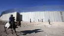 Pria Palestina dan seorang anak menunggangi keledai saat sejumlah anak bermain sepak bola di sebuah lapangan di samping tembok pemisah Israel - Palestina di desa Beit Awa di Tepi Barat yang diduduki Israel (12/11). (AFP Photo/Hazem Bader)