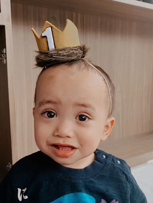 Biru terlihat sangat lucu dan menggemaskan ketika mengenakan seperti hiasan untuk kepala berbentuk mahkota. Dalam potret ini terlihat wajah Xabiru dengan senyum terpaksa tapi menggemaskan. (Liputan6.com/IG/rachelvennya)