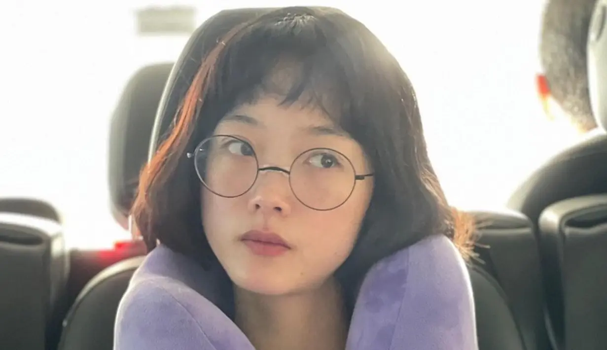 Penampilannya semakin cute dengan penggunaan kacamata frame bulat. Makeup natural yang digunakan juga memperlihatkan wajahnya yang awet muda bak masih ABG. Bergaya dengan pose berpikir dan mengenakan bantal leher ungu membuatnya jadi terlihat begitu manis. (Liputan6.com/IG/@leeyoum262)