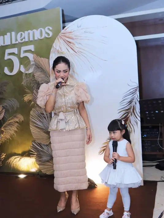 Krisdayanti tampak mengenakan busana kostum serba coklat. Dengan atasan kerah dihiasi motif gold dengan lengan balon dengan aksen bertumpuk dipadukan belt kain pita serta rok panjang fit body warna coklat dengan aksen bergaris. [@krisdayantilemos]