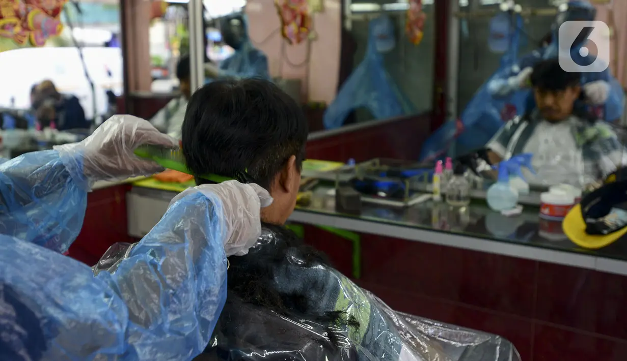 Tukang cukur mengenakan alat pelindung diri (APD) dari plastik saat mencukur rambut pelanggan di Pondok Kelapa, Jakarta, Selasa (5/5/2020). Tukang cukur mengenakan APD untuk mengurangi risiko penularan virus corona COVID-19. (merdeka.com/Imam Buhori)