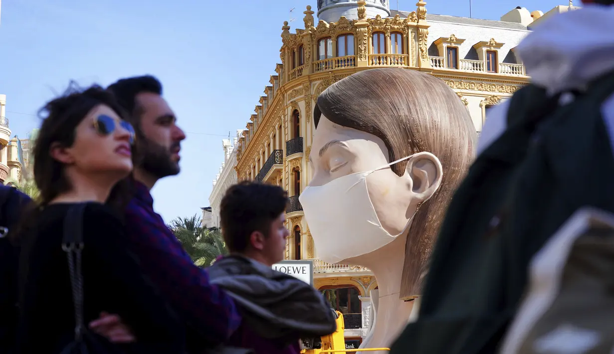 Orang-orang melintasi patung raksasa mengenakan masker yang akan ditampilkan di festival Las Fallas atau festival api di Valencia, Spanyol, Rabu (11/3/2020). Festival Fallas yang akan berlangsung pada 13 Maret telah dibatalkan karena wabah coronavirus. (AP/Alberto Saiz)