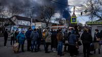 Orang-orang mengantre di depan supermarket sementara asap mengepul di atas kota Vasylkiv di luar Kiev (27/2/2022). Rudal Rusia telah menghantam pipa gas dan kilang minyak di kawasan Vasylkiv. (AFP/Dimitar Dilkof)