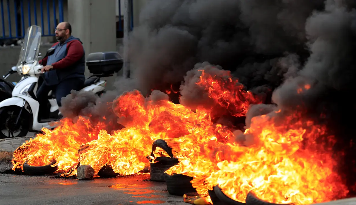 Seorang pria mengendarai skuternya melewati ban yang terbakar di salah satu jalan utama selama protes terhadap kenaikan harga barang-barang konsumsi dan jatuhnya mata uang lokal di Beirut, Lebanon, Senin (29/11/2021). (AP Photo/Hussein Malla)