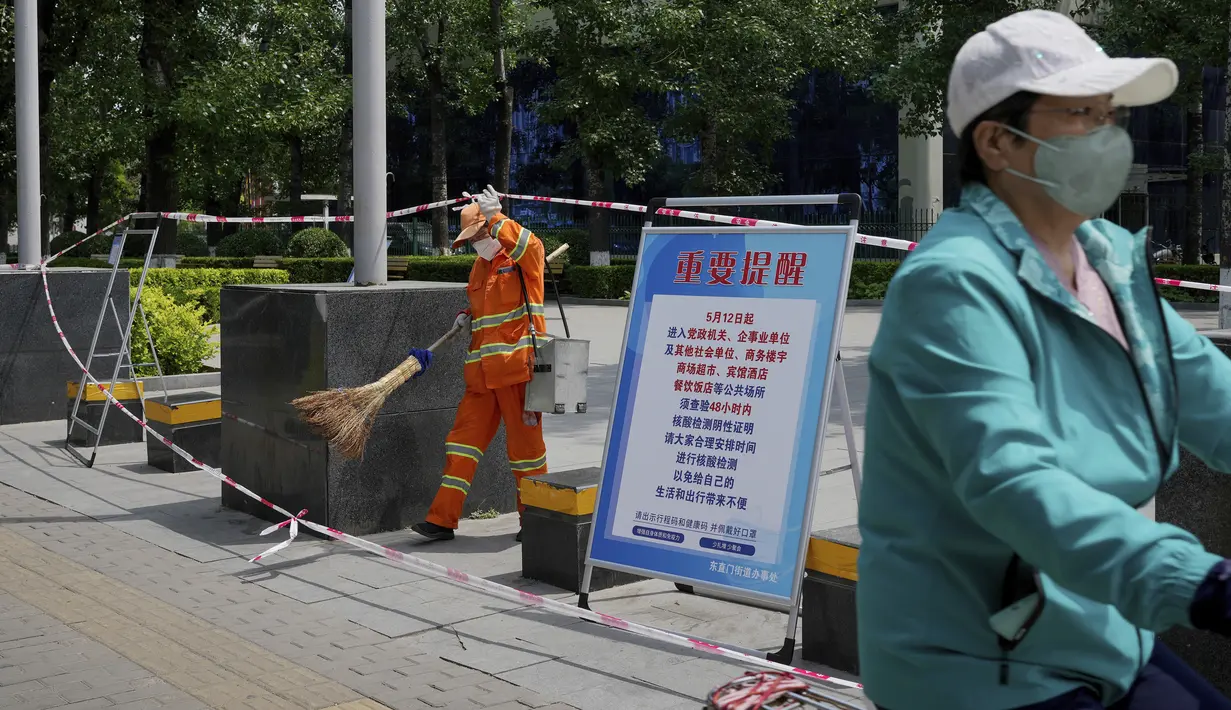 Seorang perempuan yang mengenakan masker berkendara dekat petugas kebersihan di alun-alun yang dibarikade untuk mencegah warga berkumpul karena tindakan pandemi di Beijing, China, Selasa (17/5/2022). Pada Minggu (15/5) kemarin, Beijing memperpanjang aturan untuk bekerja dari rumah di empat distrik. Itu sudah melarang layanan makan di restoran dan membatasi transportasi umum, di antara langkah-langkah lainnya. (AP Photo/Andy Wong)