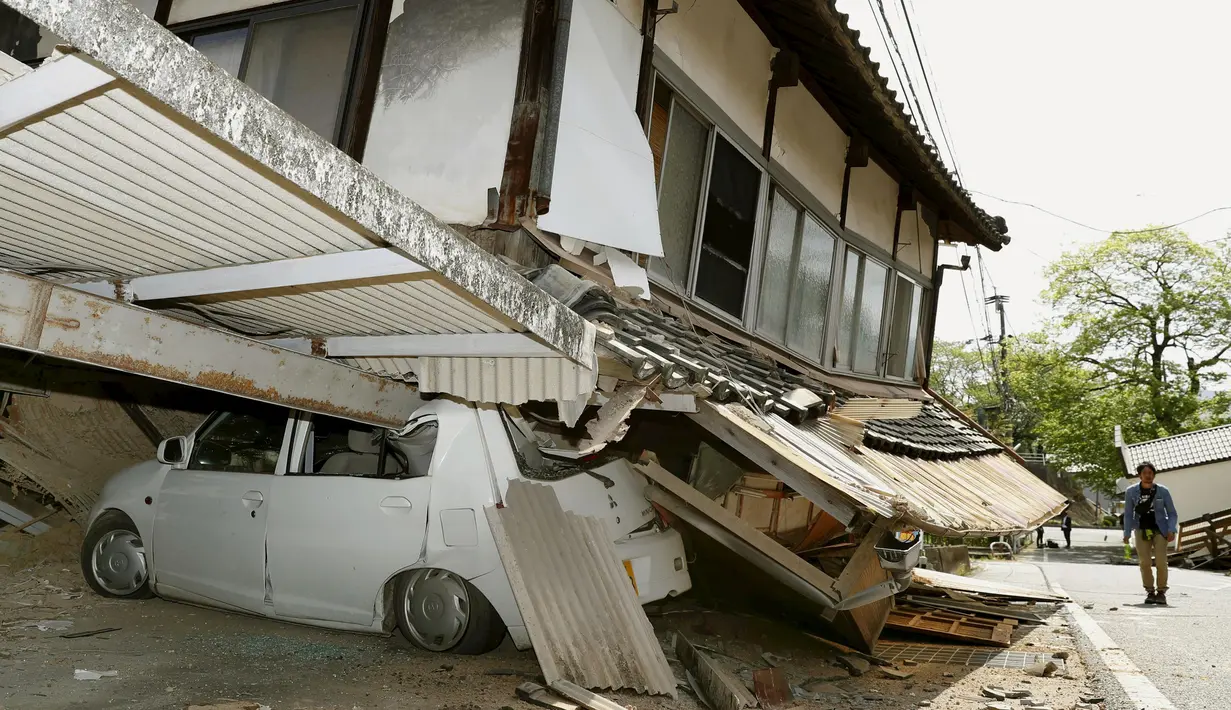 Warga berjalan di depan mobil yang tertimpa reruntuhan rumah di kota Mashiki, Kumamoto, Jepang selatan, Jumat (15/4). Gempa 6,4 SR yang mengguncang pada Kamis (14/4) waktu setempat, mengakibatkan lebih dari 20 bangunan rumah hancur. (REUTERS/Kyodo)