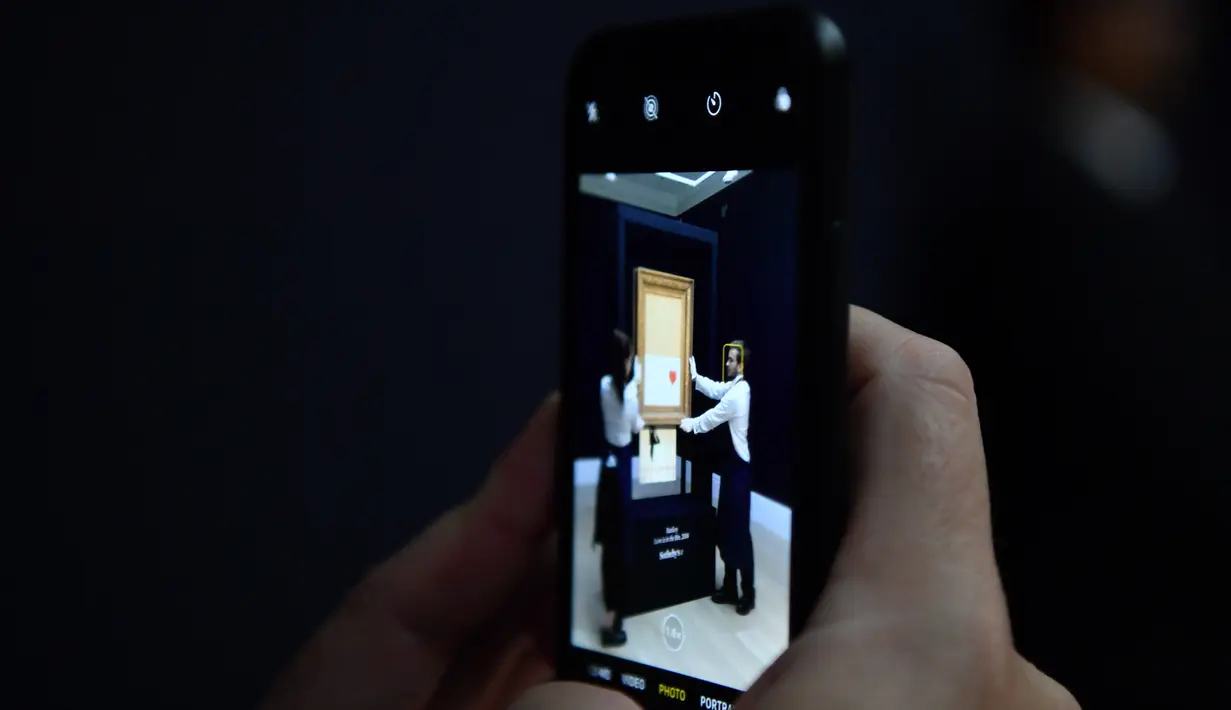 Jurnalis menggunakan smartphone mengambil gambar lukisan "Girl with Balloon" di rumah lelang Sotheby London, 12 Oktober 2018. Beberapa pengamat seni mengatakan lukisan itu kemungkinan bernilai lebih tinggi karena berubah menjadi terurai (BEN STANSALL/AFP)