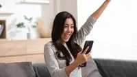 Wanita muda sedang melihat ponsel sambil tertawa dengan kabar baik (Shutterstock/220 Selfmade studo)