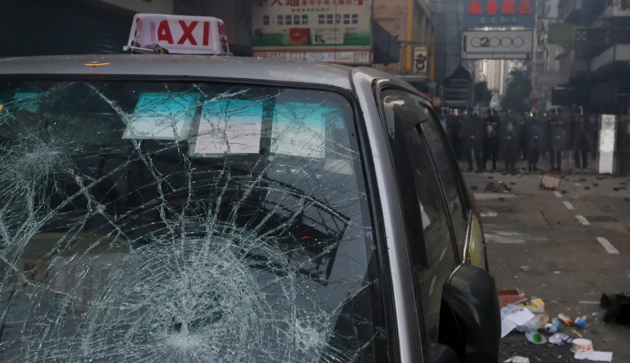 Polisi anti huru-hara berdiri di belakang taksi yang rusak akibat bentrokan dengan pengunjuk rasa di Mongkok, Hong Kong, Selasa (9/2). Bentrokan dimulai ketika pemerintah daerah mencoba untuk menertibkan PKL yang beroperasi. (REUTERS / Bobby Yip)