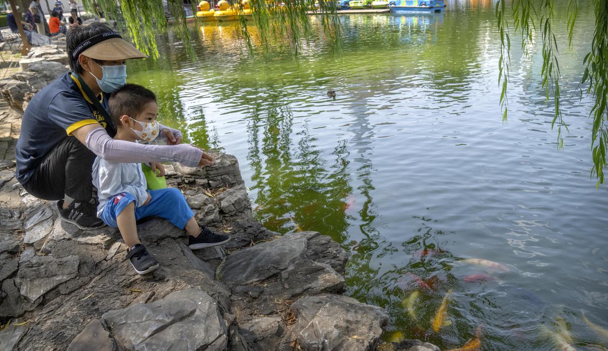 Seorang perempuan dan anak laki-laki mengenakan masker memberi makan ikan di kolam di taman umum di Beijing, Jumat (8/7/2022). Keputusan mandat vaksin Covid-19 bagi penduduk Beijing menimbulkan pro dan kontra di kalangan penduduk. Banyak dari mereka beranggapan, kebijakan itu akan sangat berdampak pada kehidupan sehari-hari. (AP Photo/Mark Schiefelbein)