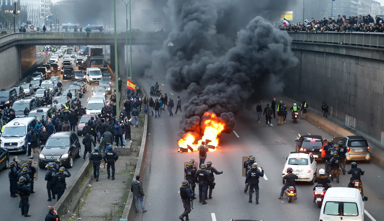Polisi anti huru-hara tiba ketika pengemudi taksi memblokir jalan dengan membakar ban saat unjuk rasa menentang layanan transportasi VTC seperti Uber, di jalan lingkar di porte Maillot di Paris, Perancis, Selasa (26/1). (AFP PHOTO/Thomas SAMSON)