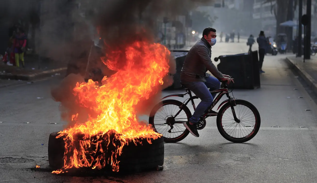 Seorang pria mengendarai sepeda melewati ban yang dibakar oleh pengunjuk rasa di sepanjang jalan utama selama protes terhadap kenaikan harga konsumen dan jatuhnya mata uang lokal di Beirut, Lebanon, Senin (29/11/2021). (AP Photo/Hussein Malla)