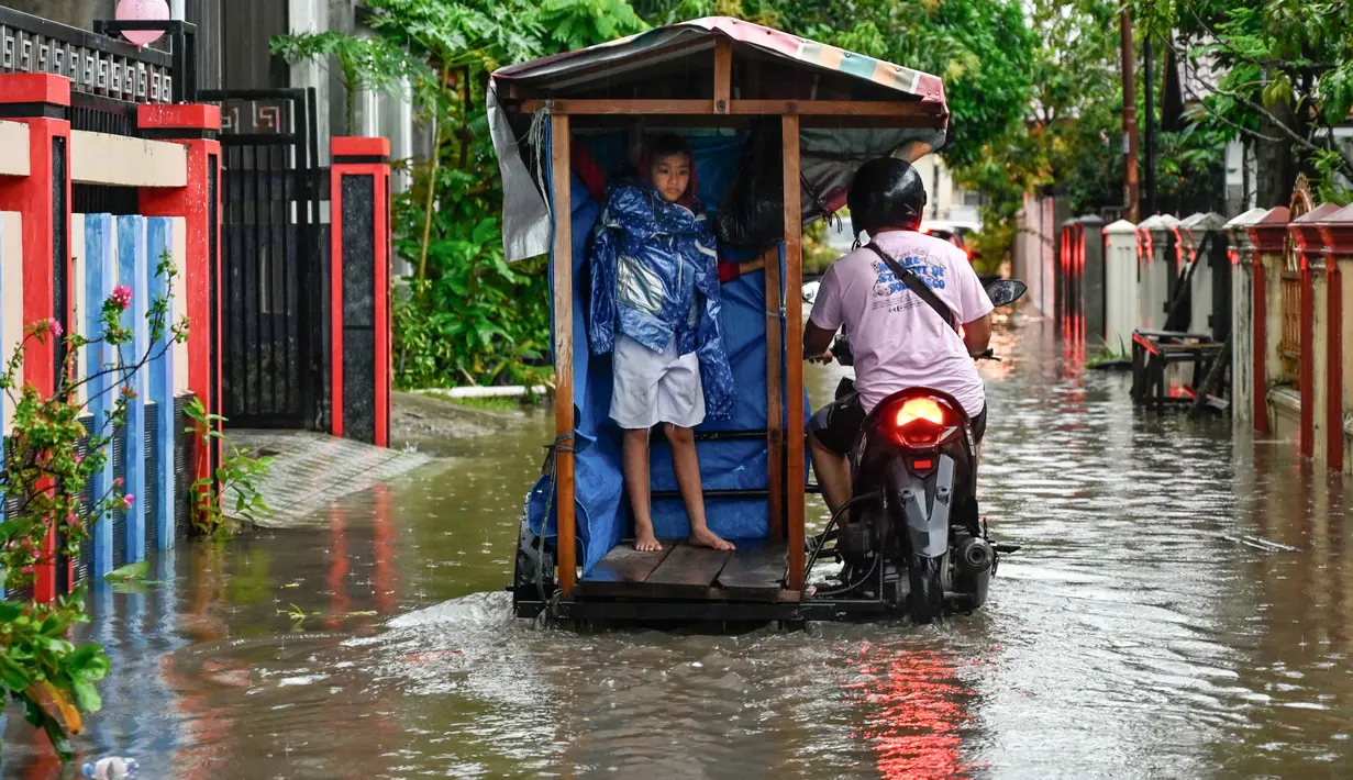 Ketinggian air bervariasi, dan di beberapa lokasi, warga terpaksa harus menembus banjir dengan sepeda motor mereka. Tampak dalam foto, seorang pria bersama seorang anak melewati banjir di jalan yang tergenang setelah hujan deras di permukiman Darul Imarah, pinggiran Banda Aceh, pada Kamis 27 November 2025. (CHAIDEER MAHYUDDIN/AFP)