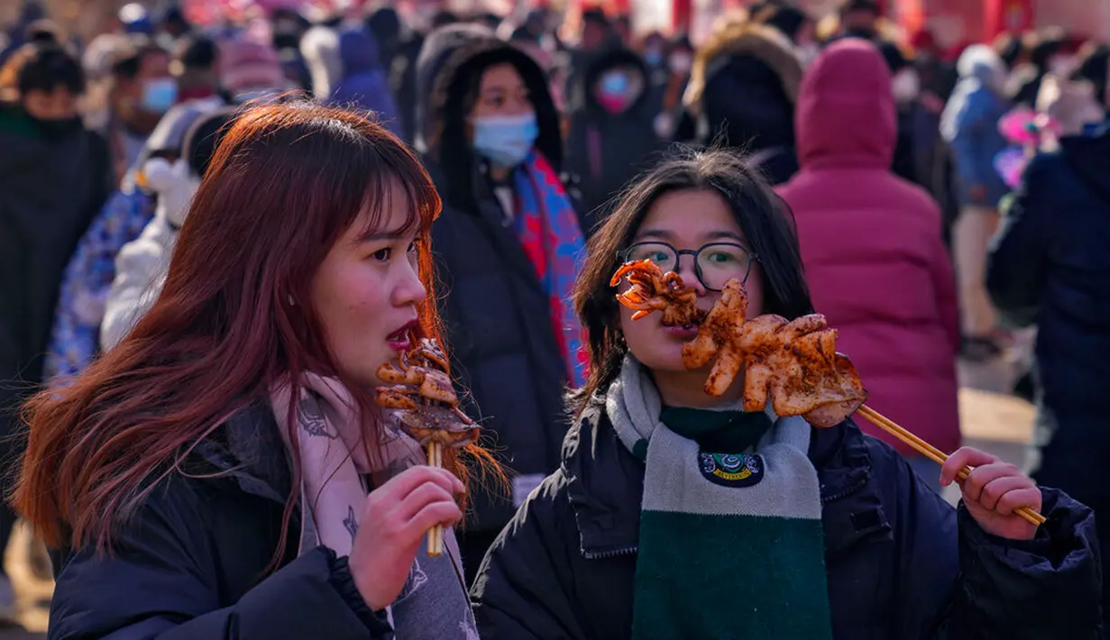 Dua wanita memakan cumi-cumi bakar saat pameran kuil pada hari kedua perayaan Tahun Baru Imlek di Taman Yuanmingyuan, Beijing, China, Senin (23/1/2023). Tahun Baru Imlek adalah hari libur tahunan terpenting di China. (AP Photo/Andy Wong)