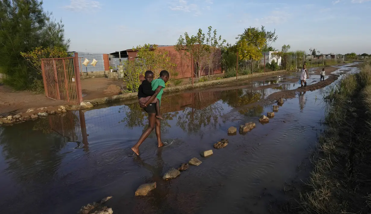 Gadis muda menggendong seorang anak di punggungnya melalui jalan yang tergenang banjir di dekat waduk yang meluap di Hamanskraal, Pretoria, Afrika Selatan, Jumat, 26 Mei 2023. (AP Photo/Themba Hadebe)