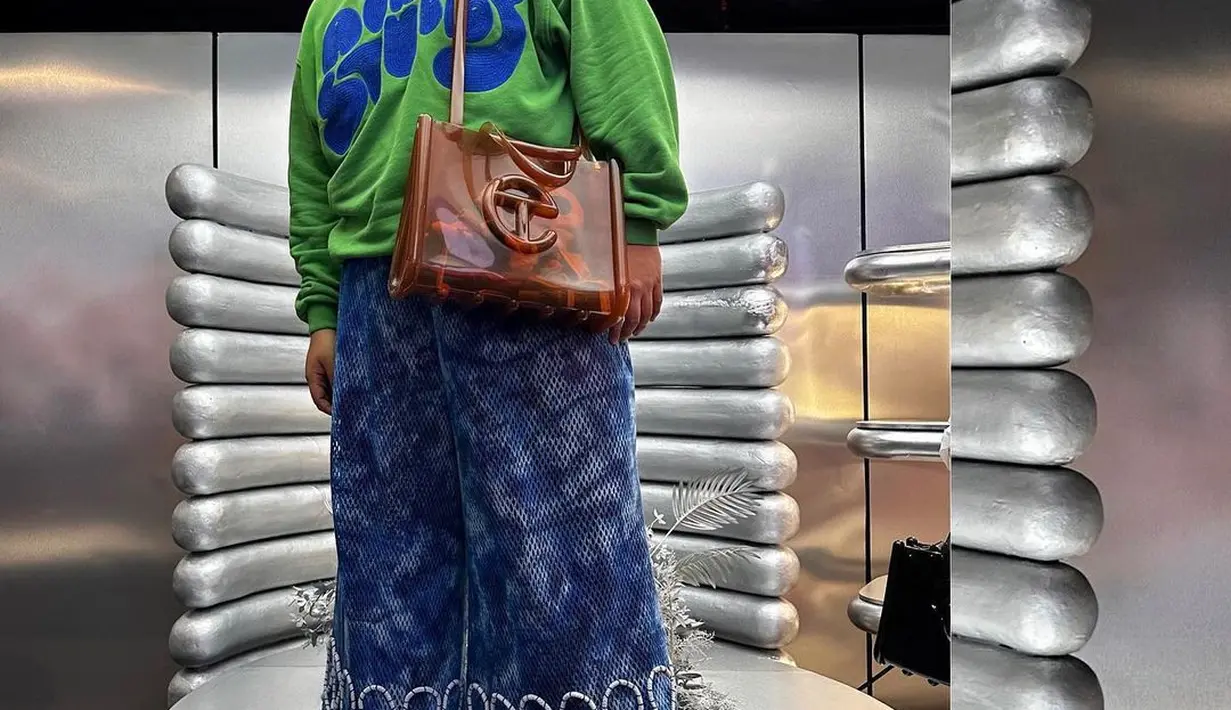 Reza Chandika tampil colorful dengan sweater hijau bermotif tulisan biru, dipadunya dengan wide leg jeans. Ia memadukan penampilannya berpose di instalasi Melissa/Telfar dengan sling totebag cokelat dari koleksi kolaborasi ini. [Foto: Instagram/rezachandika]