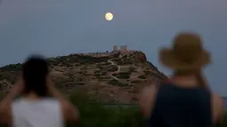 Pengunjung menyaksikan pemandangan bulan purnama hampir penuh yang menampakkan diri di langit Kuil Poseidon kuno di Cape Sounion, sekitar 70 kilometer sebelah tenggara Athena, Yunani (2/8/2020). (Xinhua/Marios Lolos)