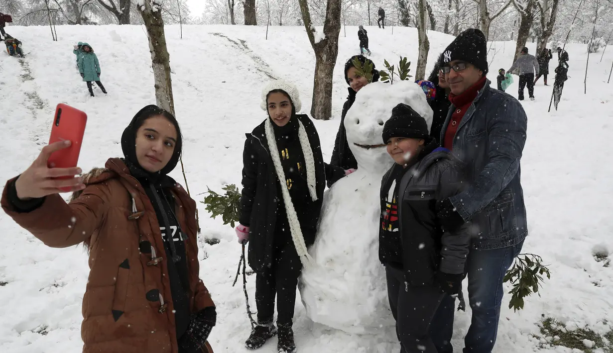 Orang-orang mengambil berswafoto dengan manusia salju ketika mereka menikmati salju di sebuah taman di Teheran utara, Iran (19/1/2020). Salju tebal menutupi jalan-jalan Teheran, menyebabkan penundaan penerbangan dan munutup sekolah, kata pihak berwenang di ibukota Iran. (AP Photo / Vahid Salemi)