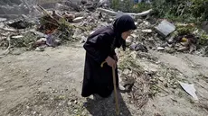 Seorang wanita lanjut usia berjalan melewati petugas pertahanan sipil Lebanon yang sedang mencari korban di antara reruntuhan, sehari setelah serangan Israel di desa Deir Qanun al-Nahr, Lebanon Selatan, pada Rabu 20 Mei 2026. (KAWNAT HAJU/AFP)