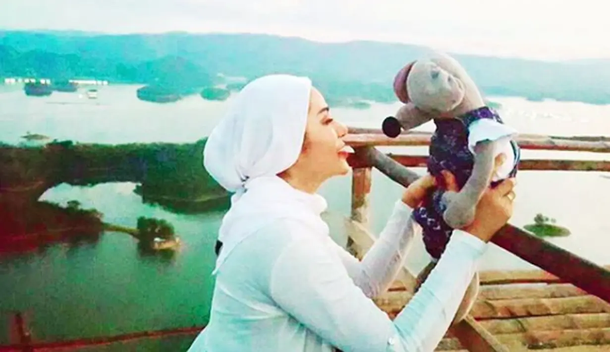 Selebritas Cinta Penelope berpose dengan sebuah boneka dengan latar belakang pemandangan alam. Banyak warga dunia maya yang memuji kecantikan Cinta Penelope lebih tampak setelah berhijab. (Instagram/princess_cinta_penelope)