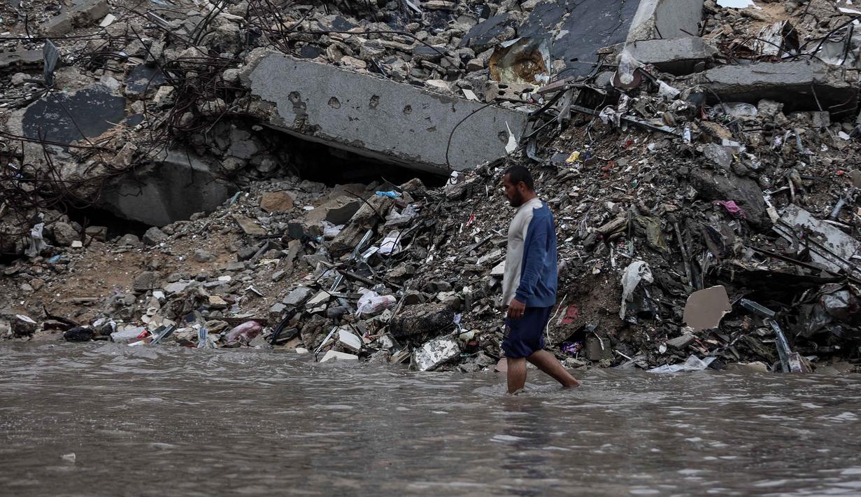Ribuan orang lainnya masih dinyatakan hilang dan diperkirakan terkubur di bawah reruntuhan. Tampak dalam foto, seorang pria berjalan di tengah banjir di kamp darurat yang menampung warga Palestina yang terlantar setelah hujan deras di Kota Gaza pada Selasa 25 November 2025. (Omar AL-QATTAA/AFP)