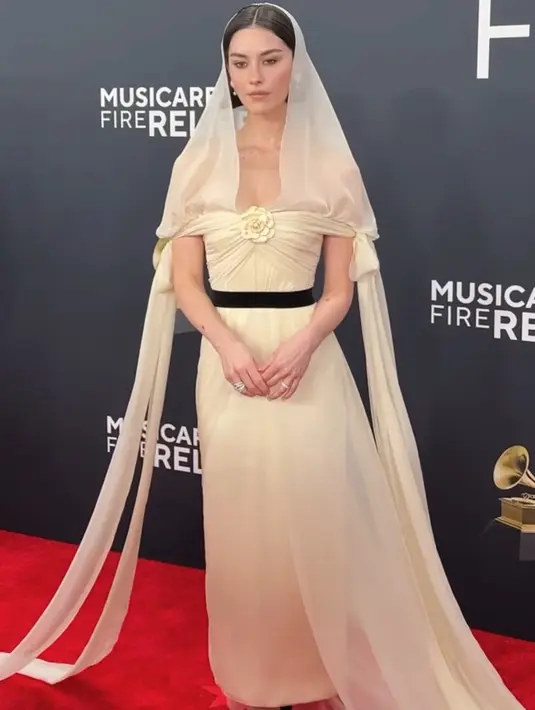 Gracie Abrams tampil dengan gaun bak pengantin warna kuning soft. Gaun dari Channel ini memiliki model off shoulder dan bunga di bagian dada serta belt hitam. Dan yang menarik tudung kepala transparan yang ia kenakan. Membuat gaun tudung ini menjadi tren. [@voguemagazine]