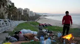 Hingga Jumat 6 Maret 2026, setidaknya ada 26 serangan udara Israel menghantam infrastruktur yang diklaim sebagai milik Hizbullah di Dahiyeh, pinggiran selatan Beirut. Tampak dalam foto, seorang pria pengungsi bersama keluarganya yang melarikan diri dari serangan udara Israel di Dahiyeh, pinggiran selatan Beirut, menyalakan api untuk menghangatkan diri di tepi pantai Beirut, Lebanon, Jumat 6 Maret 2026. (AP Photo/Hussein Malla)