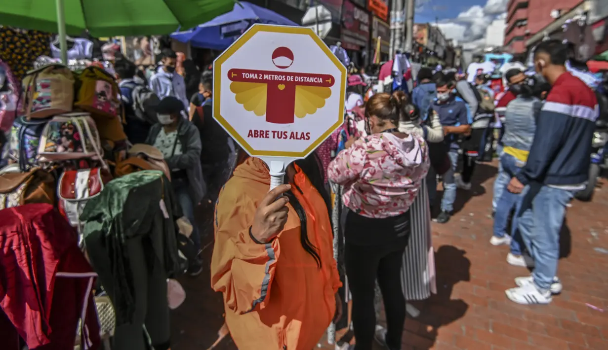 Petugas memegang tanda “Jaga jarak sejauh 2 meter” saat orang-orang berbelanja hadiah Natal di San Victorino, Bogota, Kolombia, Senin (21/12/2020). Kolombia, pada pekan lalu, kasus COVID-19 yang dikonfirmasi setiap harinya mencapai level tertinggi sejak pertengahan Agustus. (Juan BARRETO/AFP)