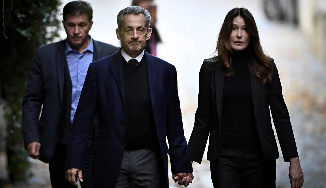 Mantan Presiden Prancis Nicolas Sarkozy (tengah), bersama istrinya Carla Bruni-Sarkozy, berjalan meninggalkan kediamannya untuk menyerahkan diri ke Penjara La Sante guna menjalani hukuman penjara lima tahun setelah dinyatakan bersalah atas konspirasi kriminal terkait rencana pendanaan kampanye pemilu 2007-nya oleh diktator Libya Moamer Kadhafi, di Paris, pada 21 Oktober 2025. (JULIEN DE ROSA/AFP)