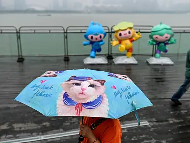 Seorang pejalan kaki menggunakan payung saat melintas di pedestrian Sungai Qiantang. Hangzhou, Zhejiang, China yang berhias maskot Asian Games 2022 (Chenchen, Congcong, dan Lianlian) pada 22 September 2023 waktu setempat. (AFP/Manan Vatsyayana)
