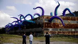 Pengunjung mengambil gambar patung karet "Sea Monsters HERE" di gudang berkarat Building 61 di Navy Yard, Philadelphia, Selasa (9/10). Kreasi sepanjang 40 kaki ini menampilkan tentakel ungu besar yang keluar dari jendela hingga ke atap. (AP/Matt Rourke)