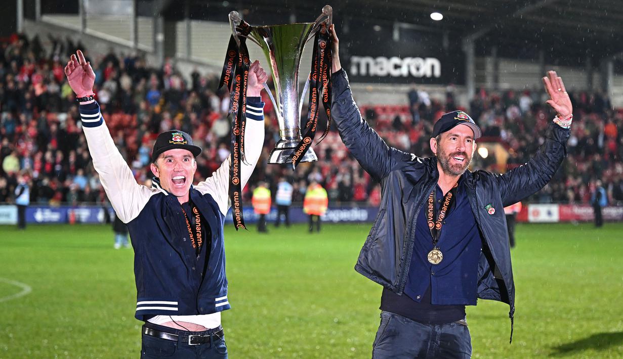 Aktor Hollywood, Ryan Reynolds (kanan) dan pemilik klub Wrexham, Rob McElhenney merayakan kesuksesan dengan mengangkat trofi setelah klub mereka berhasil promosi ke Liga Dua Inggris. Wrexham menang atas Boreham Wood dengan skor 3-1 pada laga English National League di Racecourse Ground Stadium, Wrexham, Wales, Sabtu (22/04/2023). (AFP/Oli Scarff)